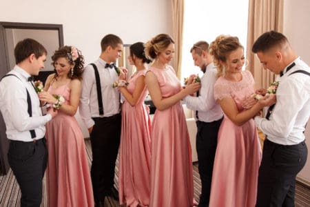 Bridesmaids in pink dresses of different styles pinning flowers on groomsmen