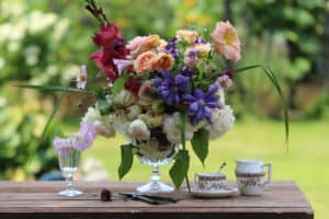 Colourful, bright fresh Bouquet of summer flowers in glass vase, china tea cup with milk jug floral pattern on aged table on fence background, garden scene, daylight, floral composition, vintage style