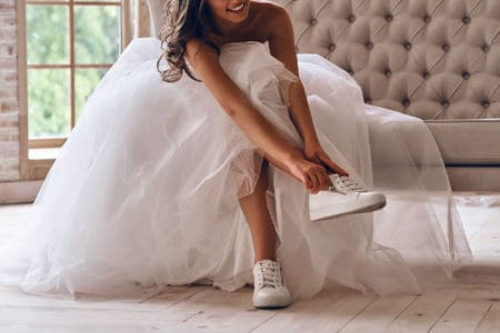 Modern bride. Attractive young woman in wedding dress putting on sports shoes and smiling while sitting on the sofa