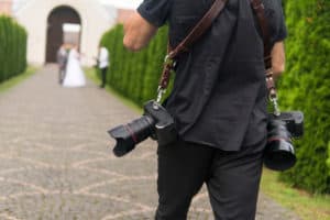 Professional wedding photographer takes pictures of the bride and groom in garden, the photographer in action with two cameras on a shoulder straps.