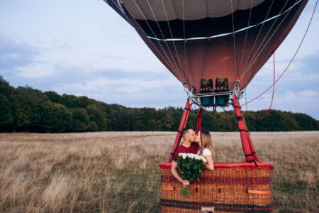 Couple in love kissing in balloon with gorgoeous bouquet of white roses on a wide field. Handsome man confessed in love to his beautiful girlfriend in air balloon flight Original proposal of marriage